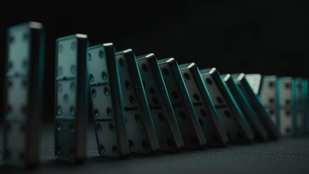 검은 공간에 늘어선 키 큰 도미노들이 차례로 넘어지는 순간. 각 블록의 모서리를 따라 청록빛이 흐른다.